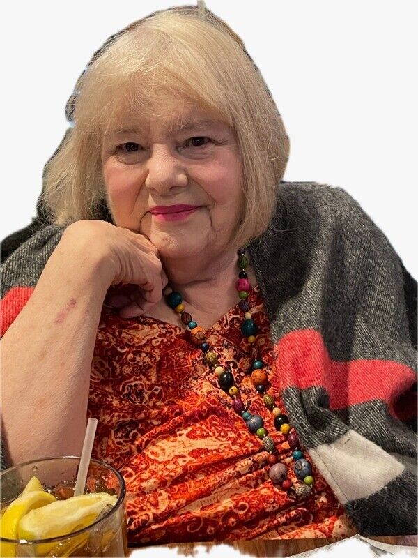 Barbara Ballard, Grandma Barb, smiling with her chin resting on her hand, wearing an orange patterned top, a colorful beaded necklace and a grey wrap